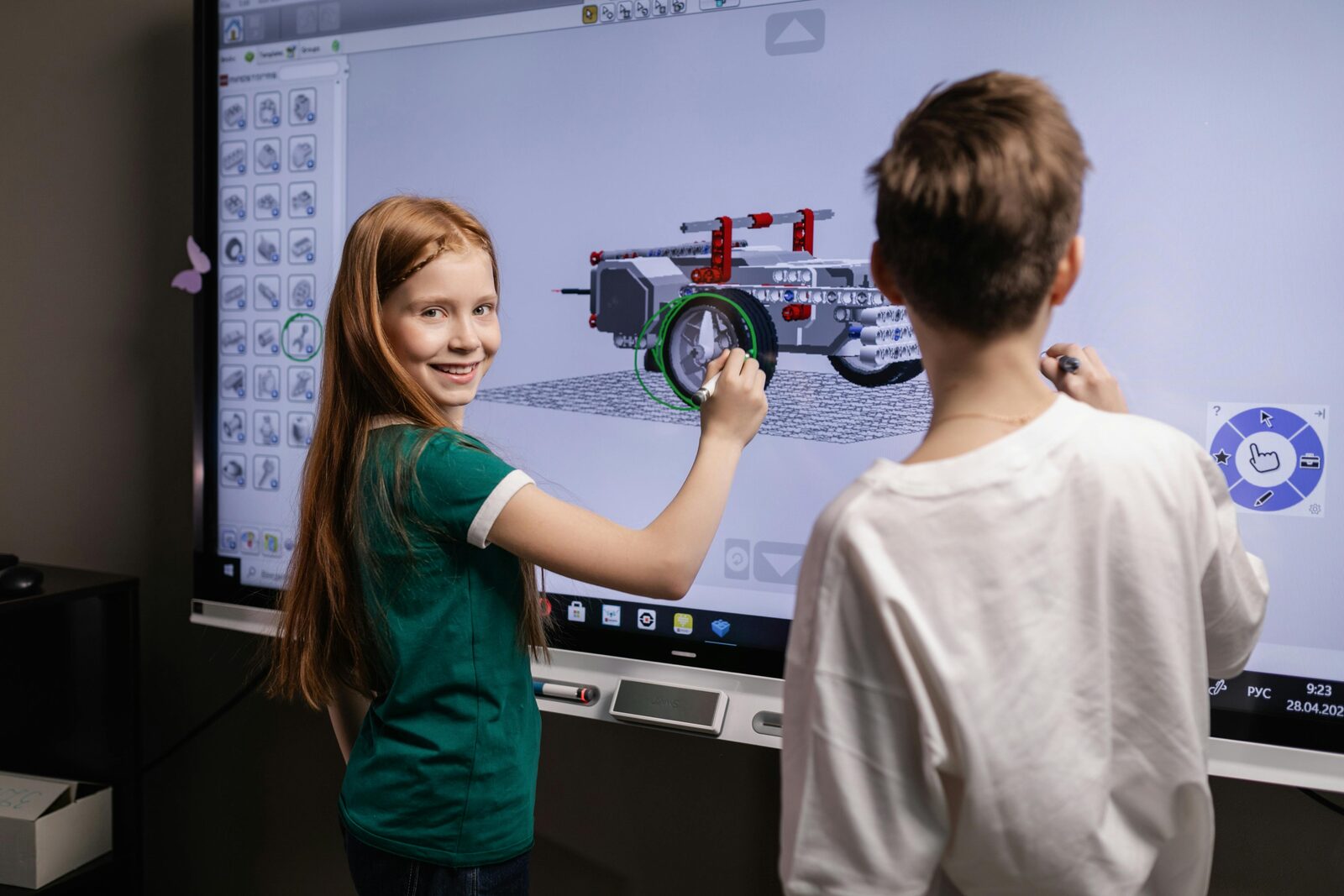 Mergaitė ir berniukas robotikos būrelyje interaktyviame ekrane konstruoja roboto modelį